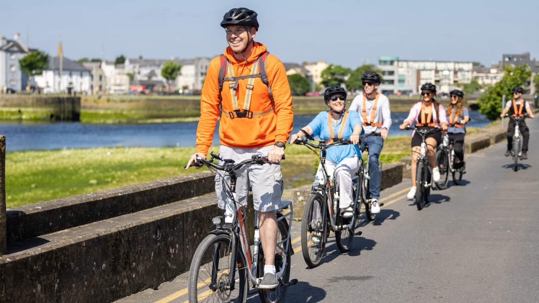Galway E-Bike Tours: Explore the City and Coast with a Local Guide A guide and a group riding electric bicycles beside Galway’s waterfront